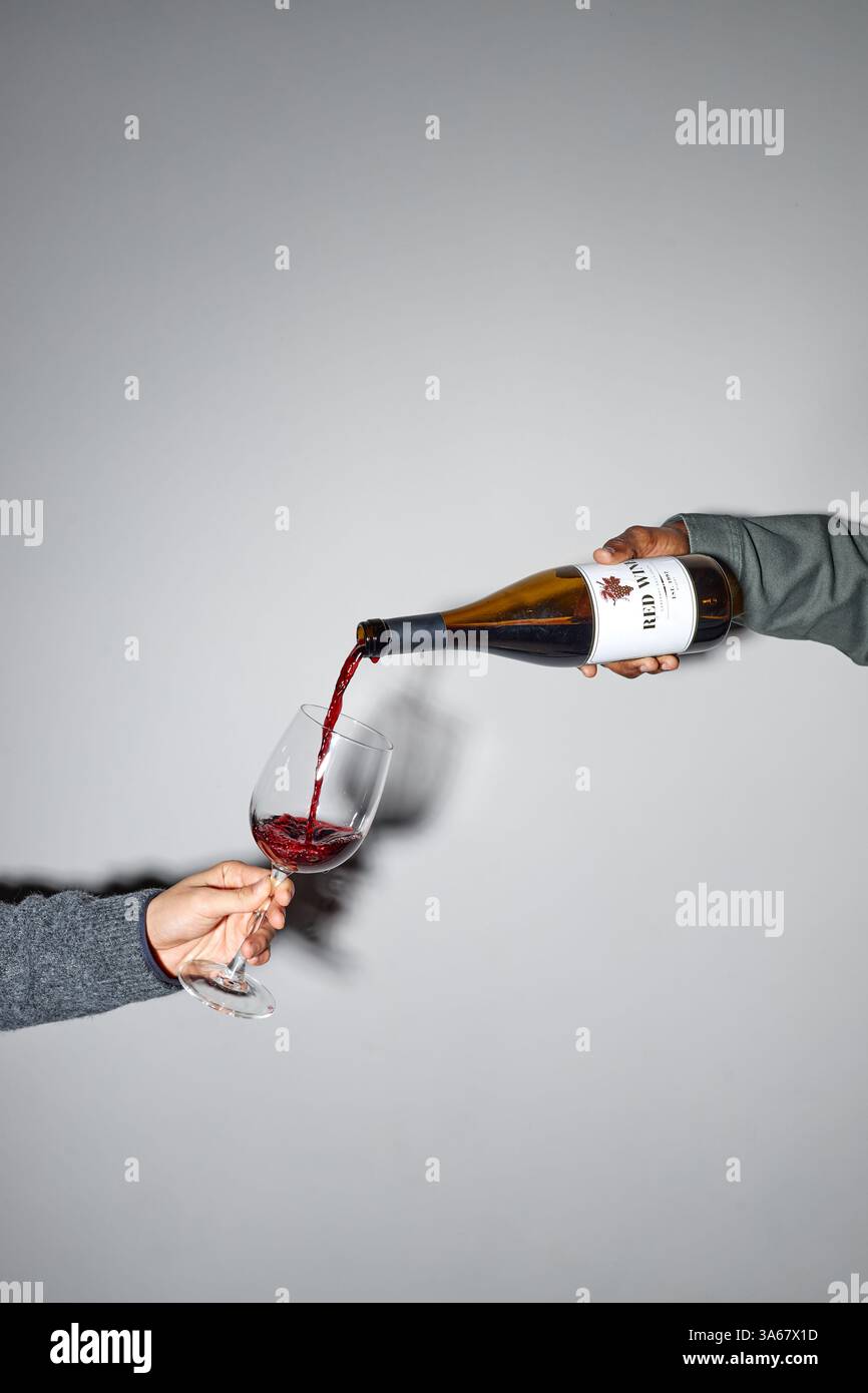 Hand gesehen beim Gießen von Rotwein aus der Flasche in Weinglas vor einem einfachen Hintergrund. Minimalistische Komposition, die die Eleganz der einfachen Aktion ohne zusätzliche Elemente betont Stockfoto