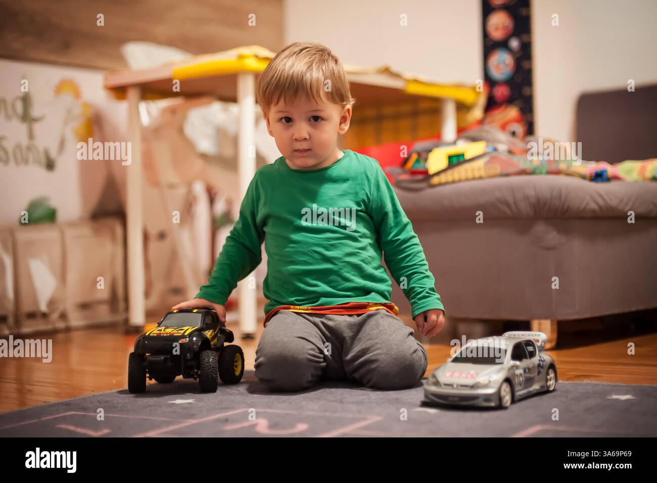 Ein kleiner Junge kniet auf einer weichen Matte im Wohnzimmer und spielt mit zwei Spielzeugautos. Das gemütliche Ambiente bietet ein komfortables Sofa und farbenfrohe Spielzeuge Stockfoto