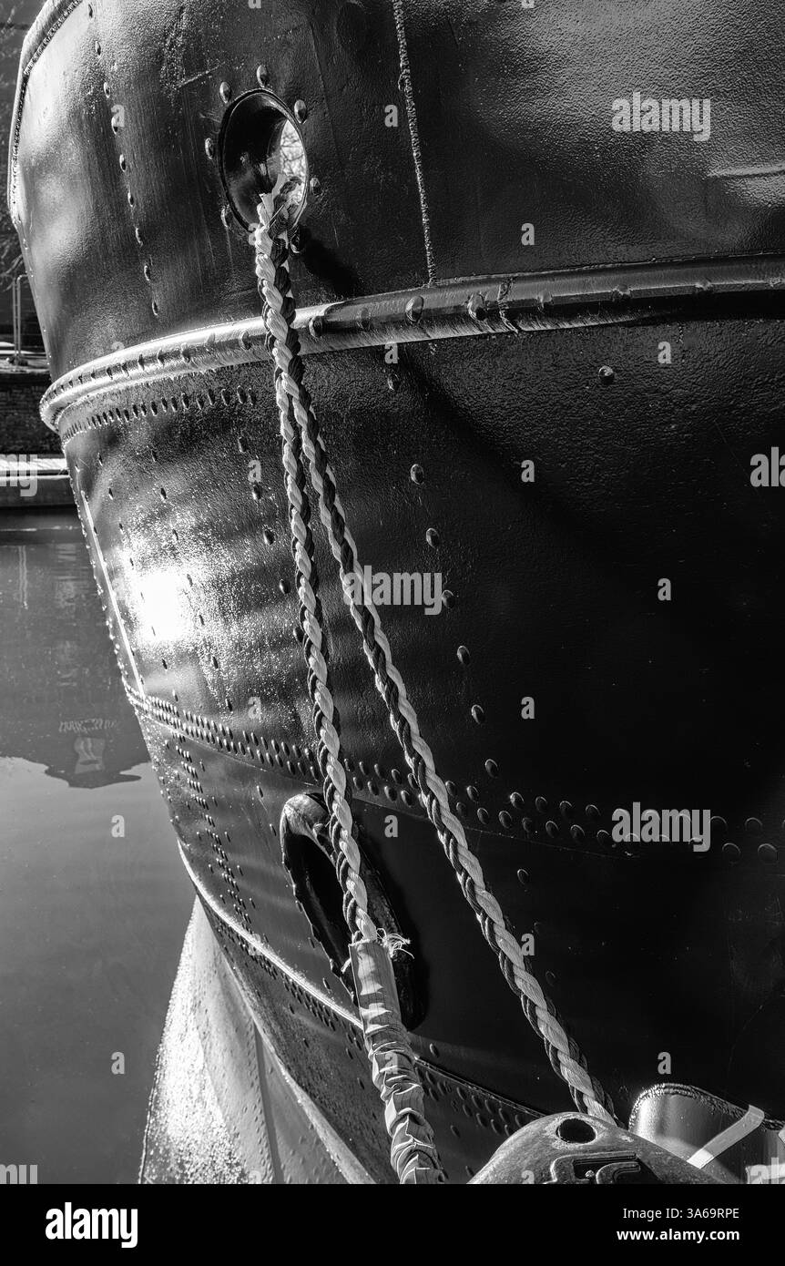 Ein Schiff ist mit einem Seil an einem Kai vertäut. Die Sonne scheint auf den Rumpf und das Schiff spiegelt sich im Wasser darunter. Stockfoto