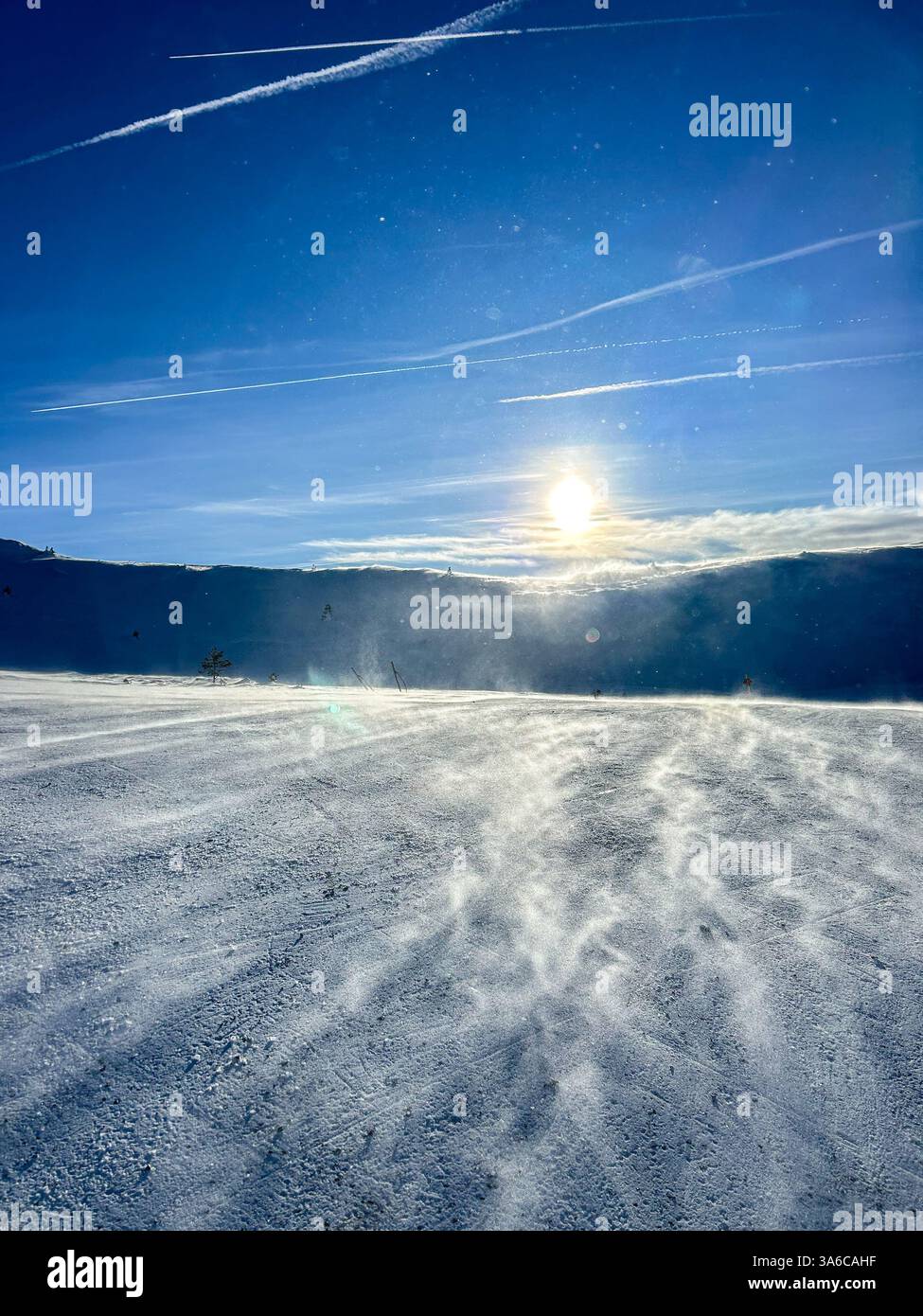 Sonnenlicht leuchtet auf Schnee, der vom Wind über eine Skipiste getragen wird, und erzeugt eine dynamische Winterszene voller Bewegung und Funkeln. - Smartphone-aufgenommenes Stockfoto