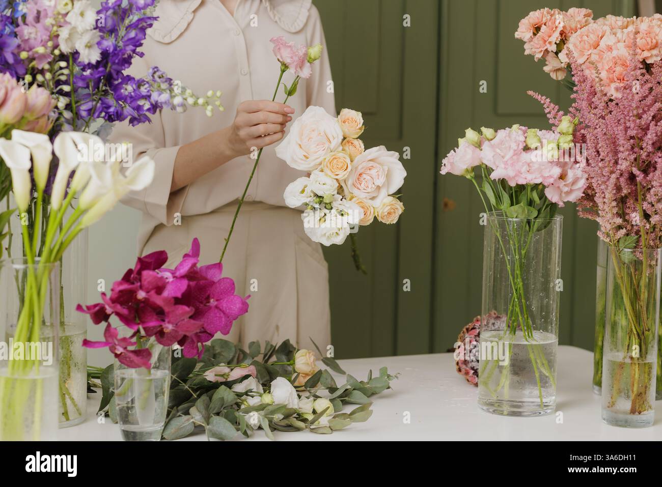 Blumenläuferin kreiert einen zarten Blumenstrauß mit frischen Blumen in einer Blumenwerkstatt, umgeben von bunten Blüten in Glasvasen. Blumenladen Stockfoto