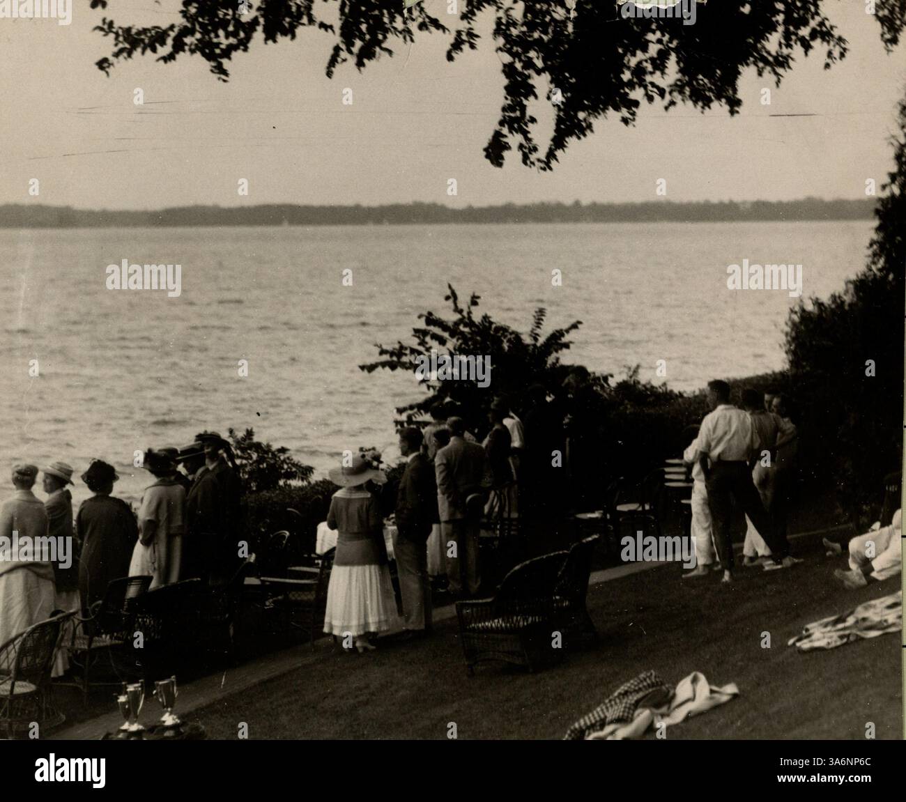 Die Gäste des E.L. Carpenter Estate am Lake Minnetonka treffen sich am Wasser, um ein Bootsrennen zu beobachten. Trophäenbecher sind im Vordergrund zu sehen. Stockfoto