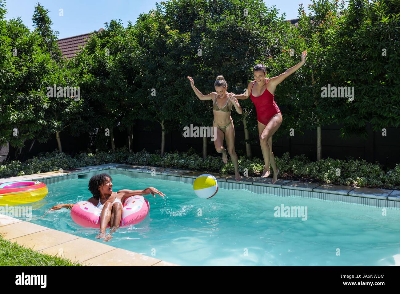 Verschiedene weibliche Freunde springen mit Strandball in den Pool, während eine andere sich im Schwimmen, Kopierraum entspannt Stockfoto