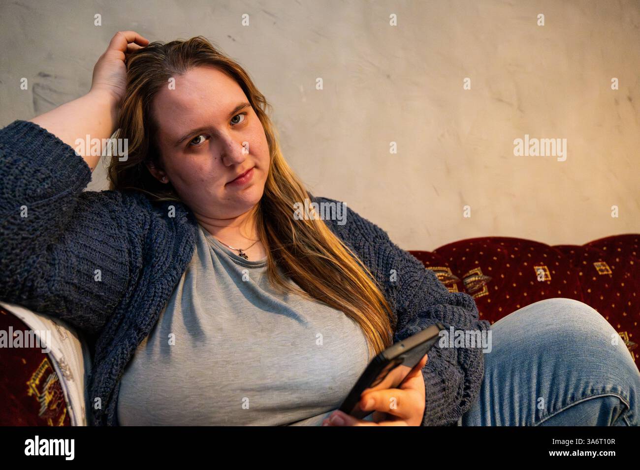 Eine entspannte Mutter in lässiger Kleidung sitzt auf einer Couch und blättert in einer ruhigen Innenumgebung durchdacht durch ihr Smartphone. Stockfoto