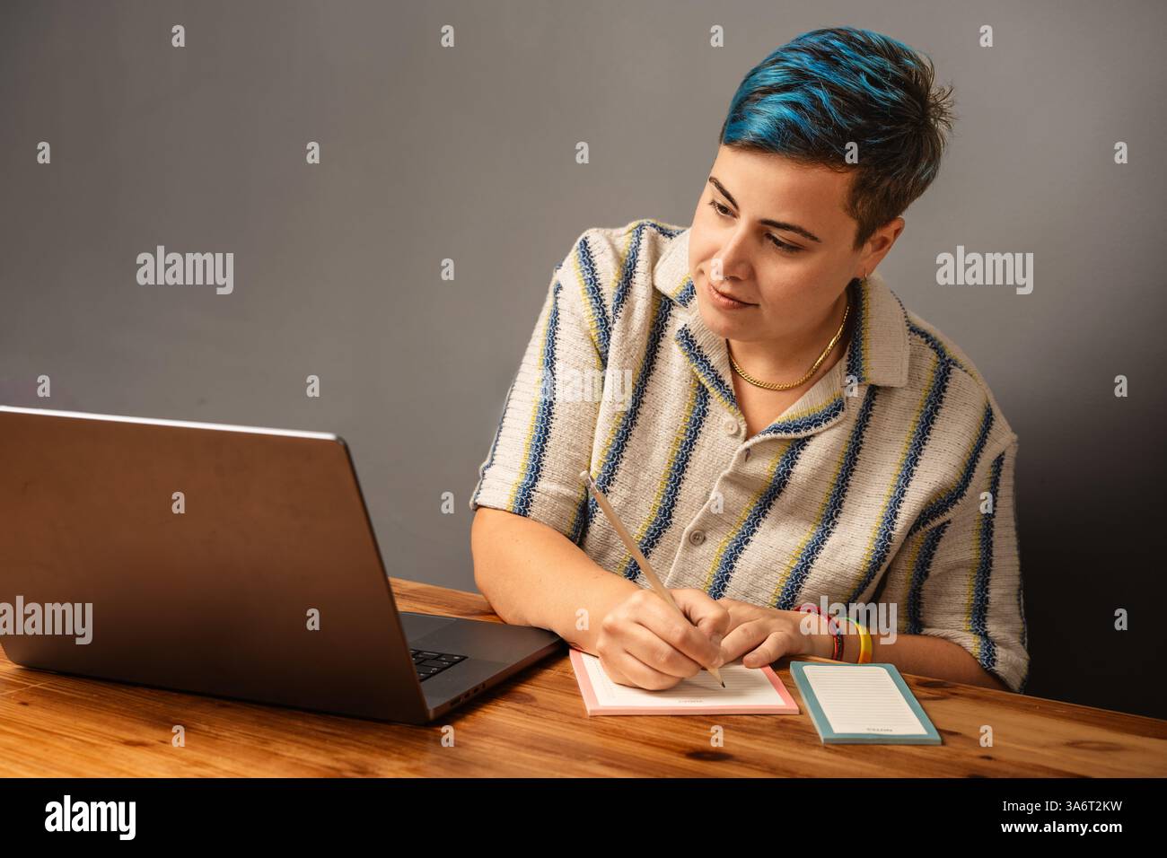 Lächelnde junge Frau mit kurzen blauen Haaren, die Notizen macht, während sie an einem hölzernen Schreibtisch auf ihrem Laptop an einem Videoanruf teilnimmt. Fernarbeit und digitale Planung Co Stockfoto