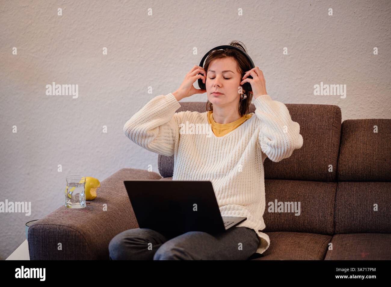 Eine Frau in einem gemütlichen Pullover sitzt auf einer Couch und passt ihre Kopfhörer an, während sie einen Laptop benutzt. Ein Glas Wasser und ein Apfel ruhen neben ihr und schaffen ein Re Stockfoto