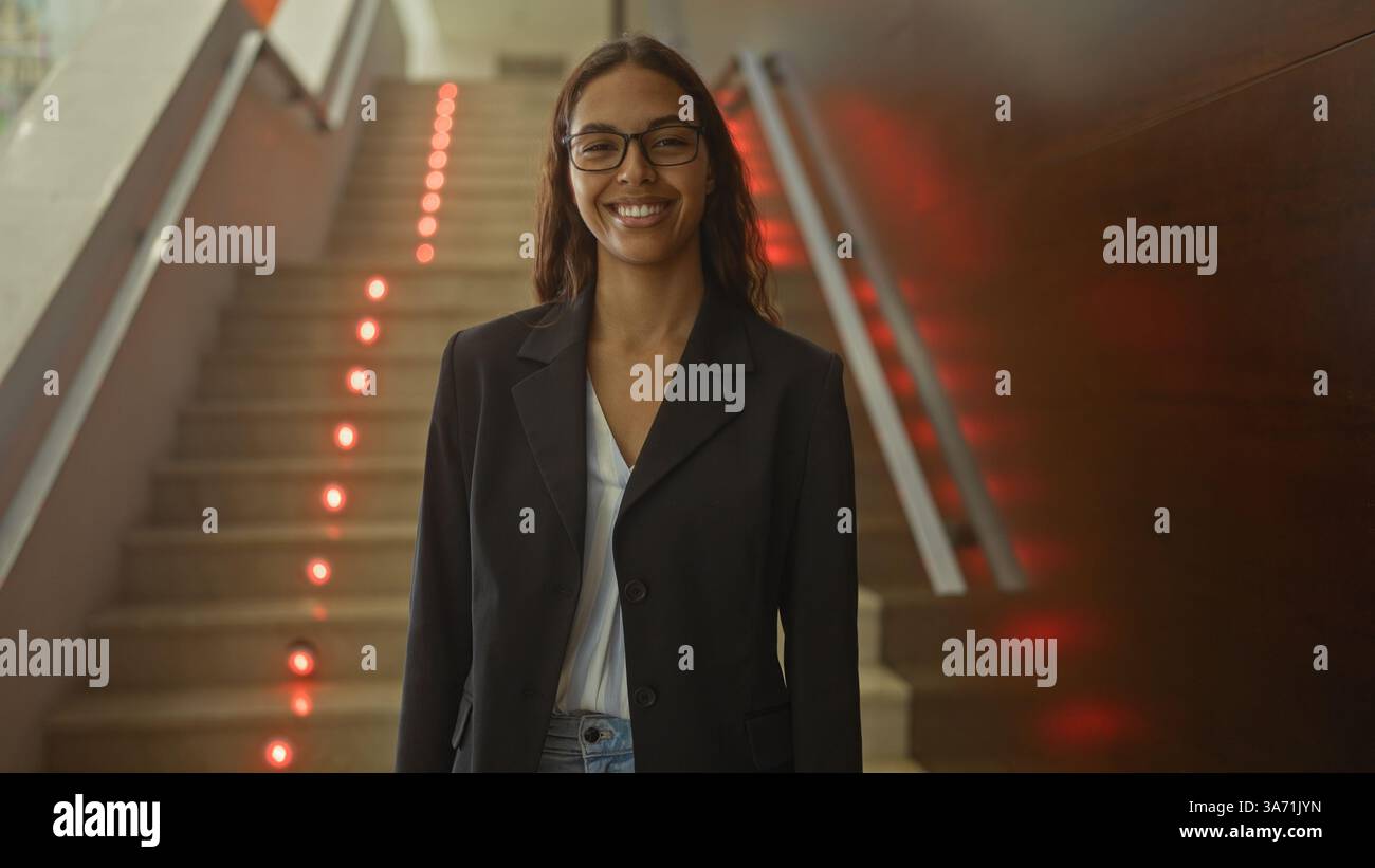 Junge hispanische Frau, die drinnen in der Nähe einer Treppe in einem Lobbykorridor steht Stockfoto