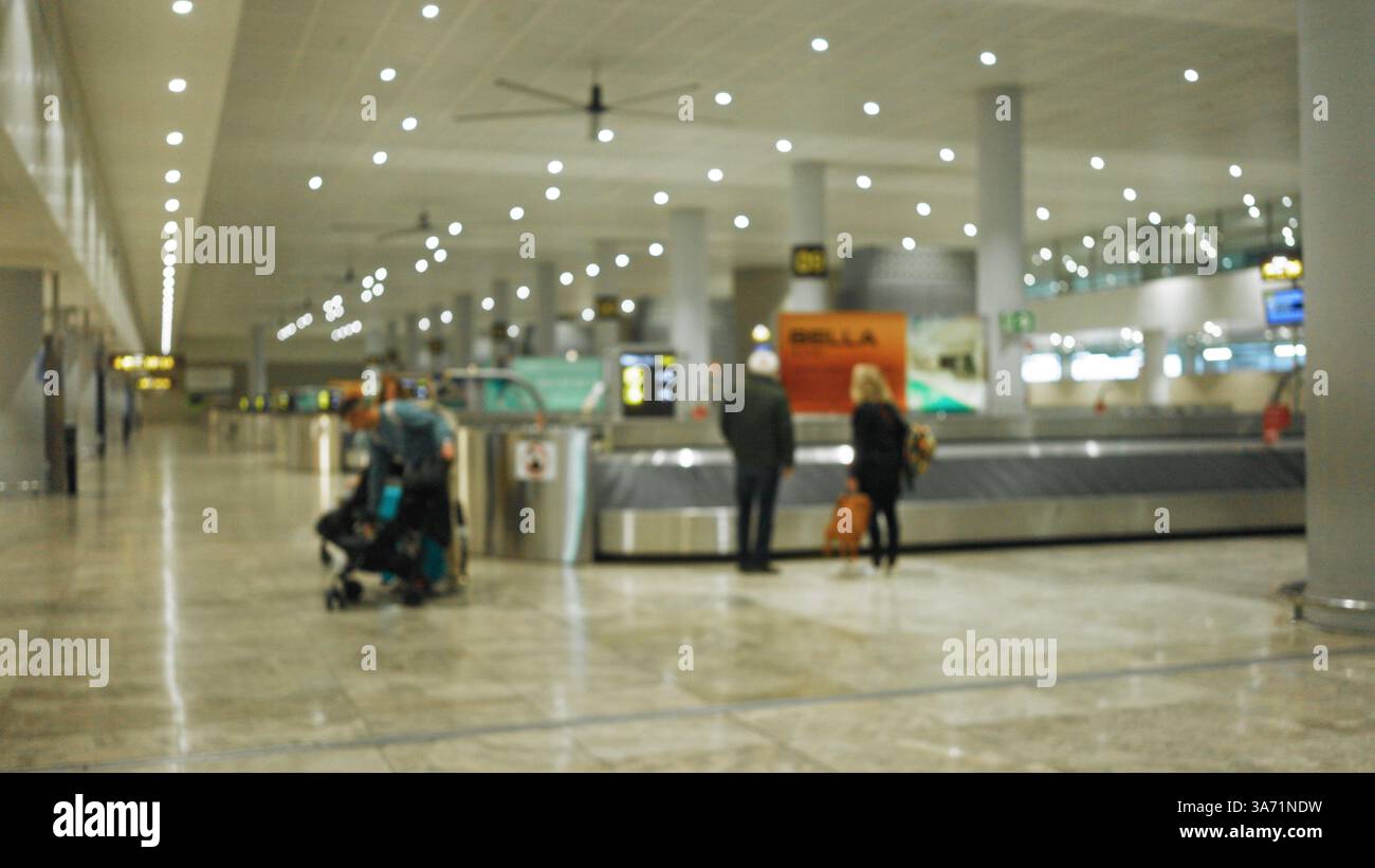 Verschwommene Reisende mit Gepäck, das an einer Gepäckausgabe im Innenbereich des Flughafens wartet, mit heller Deckenbeleuchtung und geräumigem Terminal-Design Stockfoto