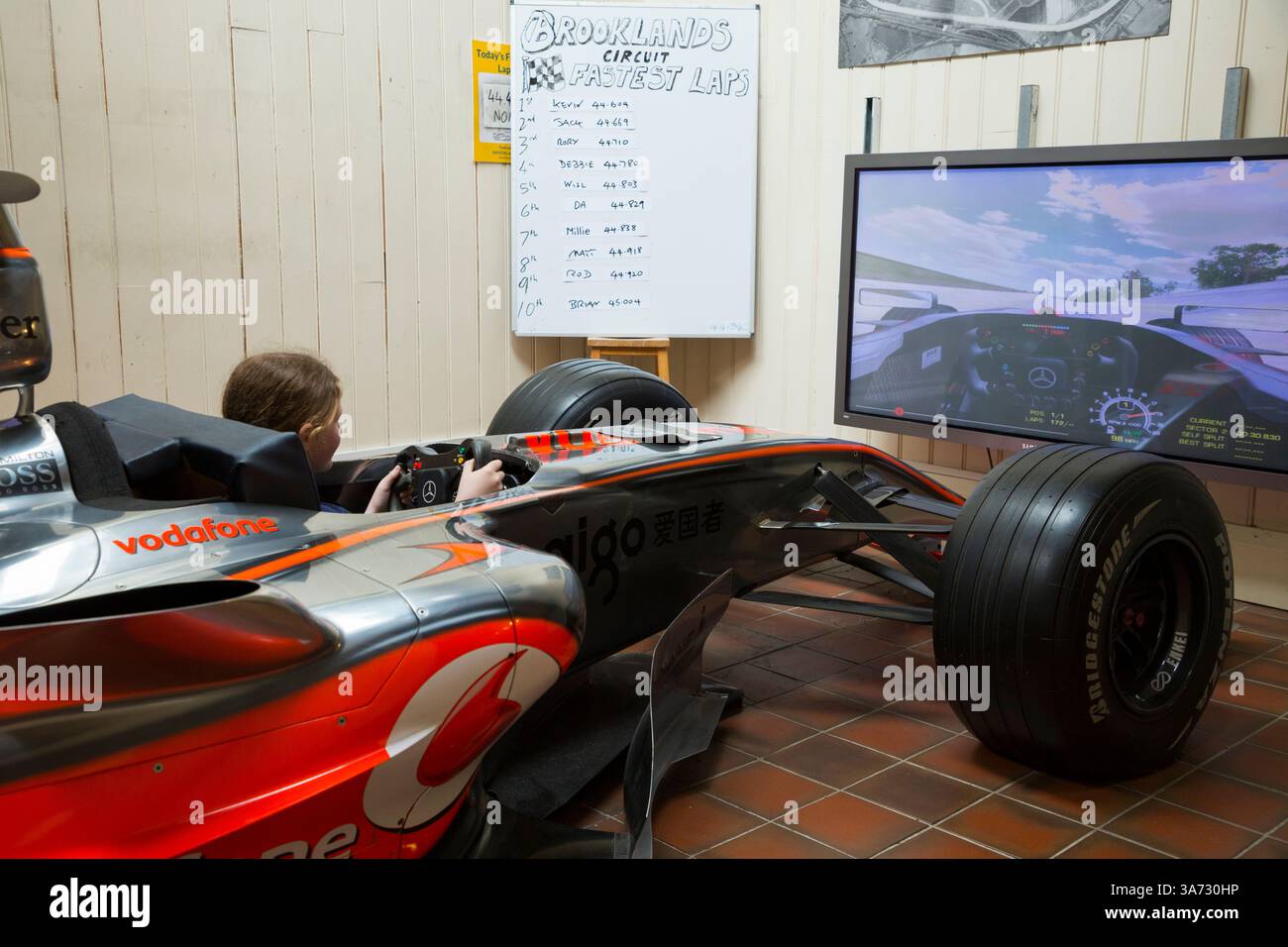 Ein kleines Mädchen im Rennwagen-Simulator ermöglicht es Besuchern, in einem MCLAREN MP4-21 F1-Showcar zu sitzen und zu fahren, um die alte Brooklands-Rennstrecke auf einem Bildschirm des Brooklands-Museums in Surrey zu erkunden. UK. (139) Stockfoto