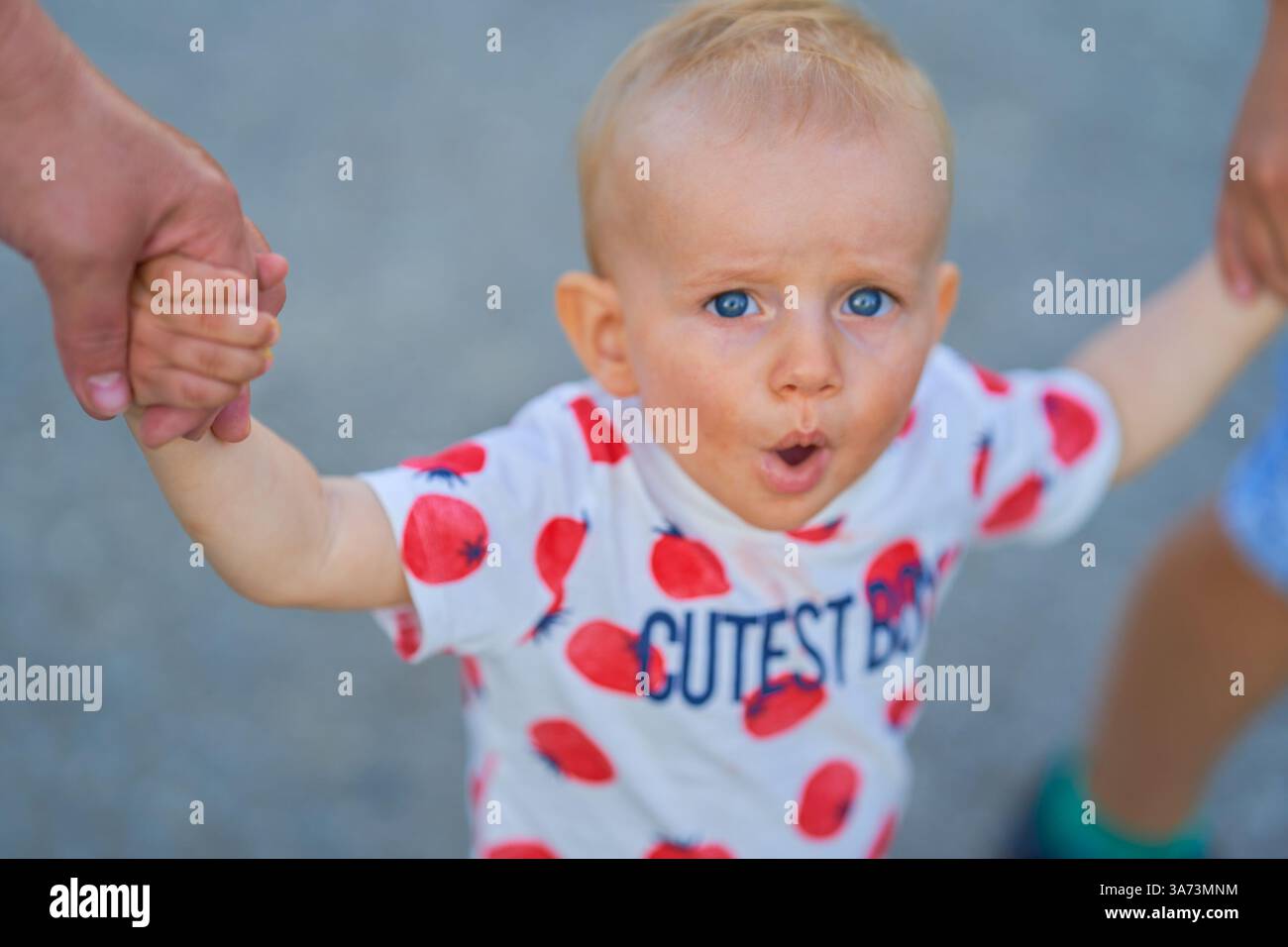 Porträt eines einjährigen Jungen, der in die Kamera blickt, blaue Augen, europäisches Aussehen mit offenem Mund, und er wird von seinen Armen an den Seiten gehalten Stockfoto