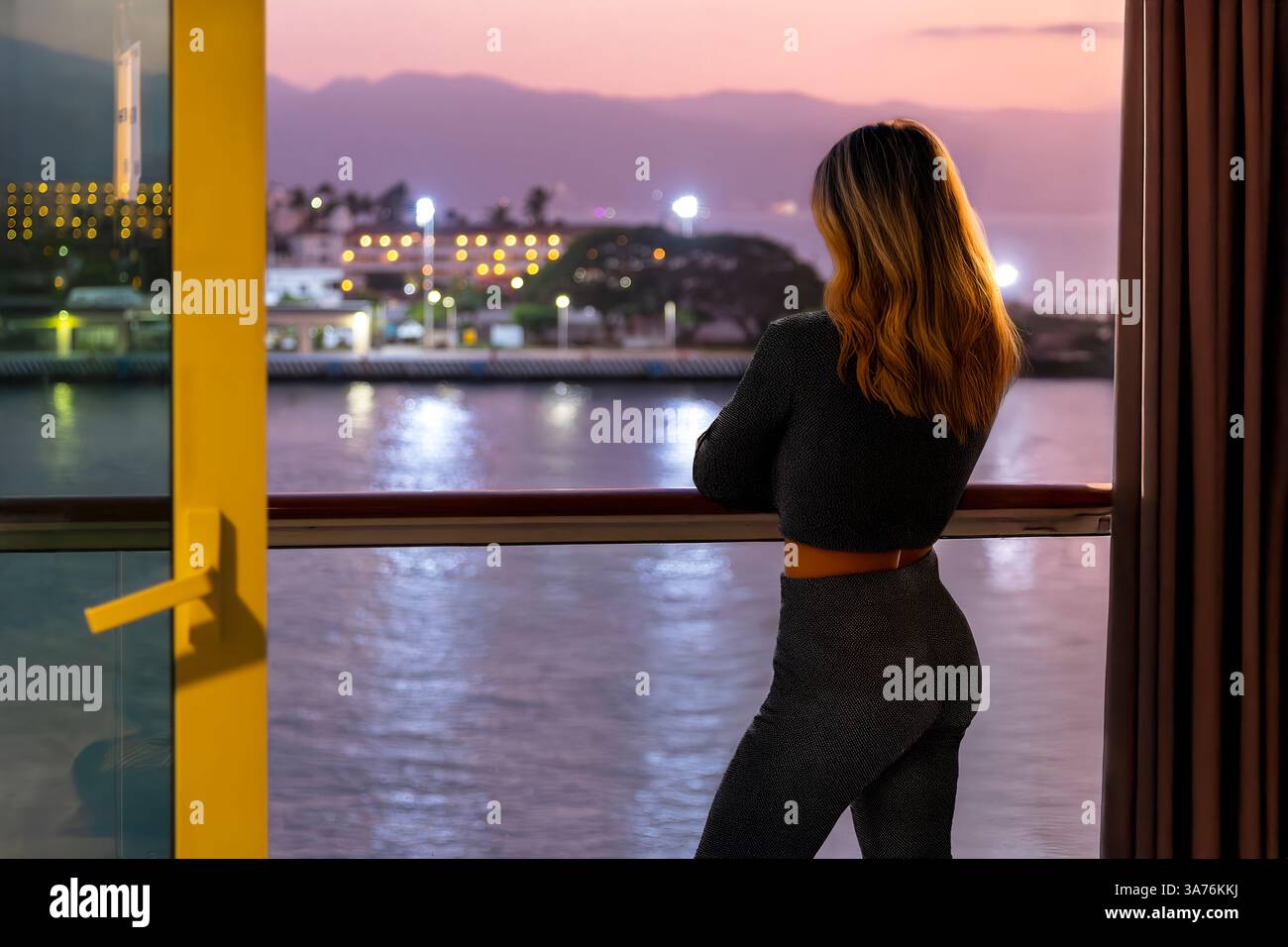 Eine junge Frau steht auf dem Balkon einer Kreuzfahrtkabine mit Blick auf den Hafen und die Skyline von Puerto Vallarta, Mexiko bei Nacht. Stockfoto