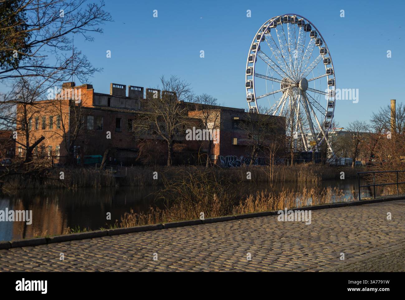 Das moderne AmberSky-Riesenrad ragt über Industriebauten und Wasser in Gdańsk, Polen. Urbane Kontraste. Stockfoto