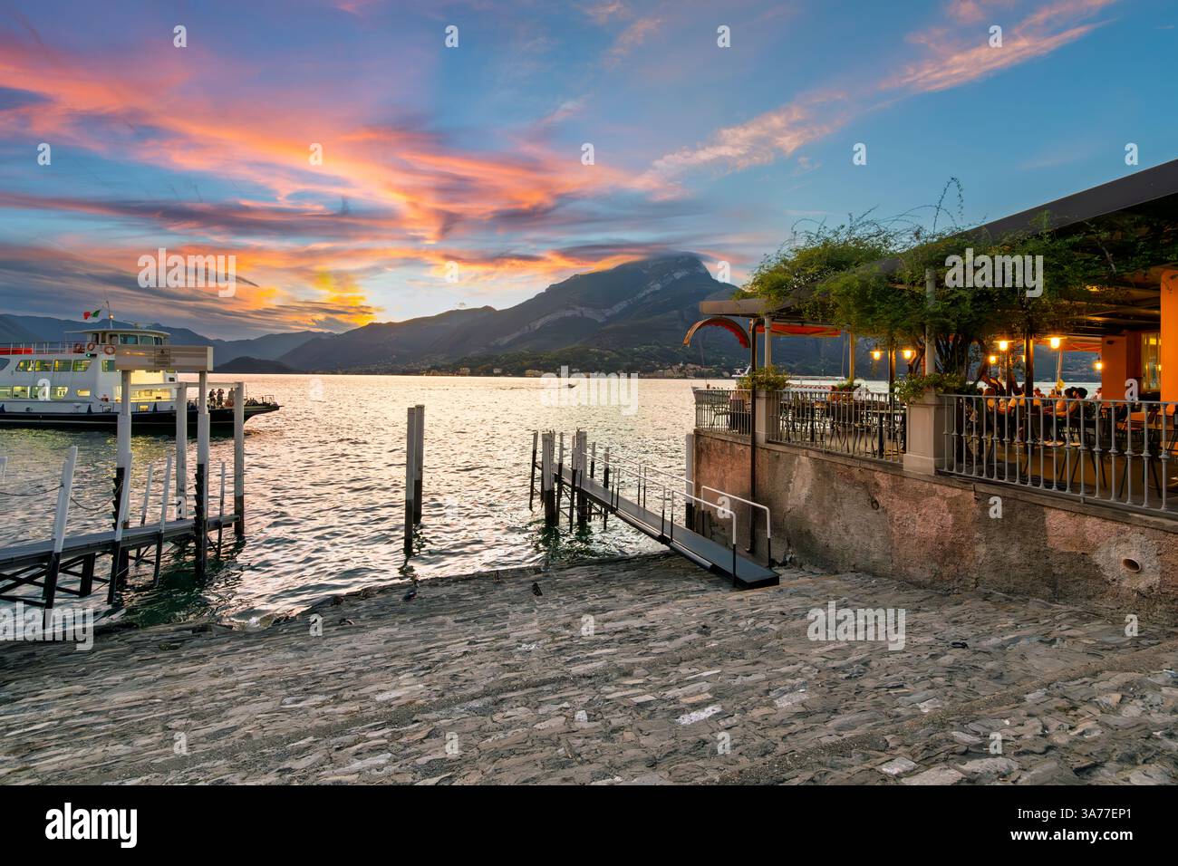 Ein Restaurant am Wasser entlang der Promenade Lungolago Europa, während die Sonne untergeht und der Himmel rosa über den Bergen und dem Comer See, Bellagio, Italien, wird. Stockfoto