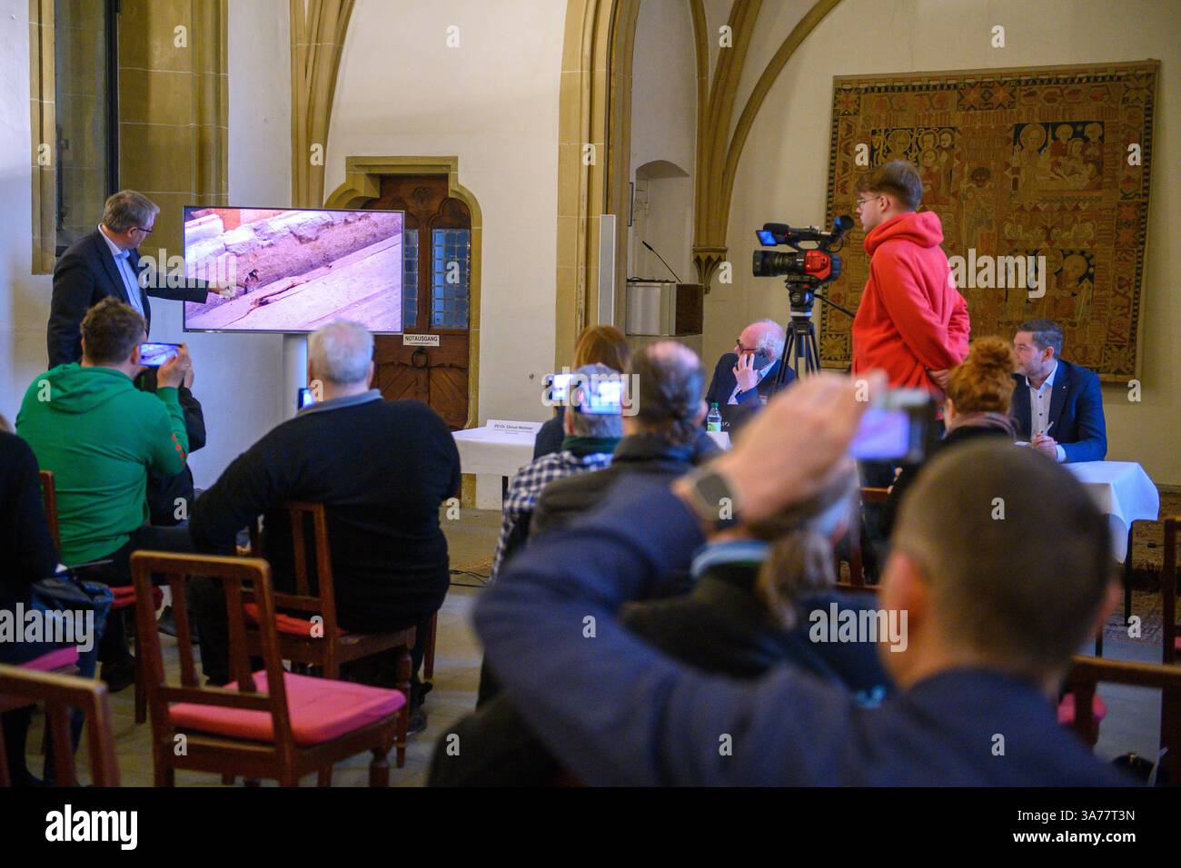 26. März 2025, Sachsen-Anhalt, Magdeburg: In der Sakristei des Magdeburger Doms erklärt Donat Wehner (l) vom Landesamt für Denkmalpflege und Archäologie Sachsen-Anhalt Medienvertretern die Beschädigung der Holzkiste im offenen Sarkophag mit Hilfe einer Live-Videoübertragung aus dem Gehege rund um das Grab Ottos des Großen. Seit Januar führen das Landesamt für Denkmalpflege und Archäologie und die Kulturstiftung Sachsen-Anhalt Untersuchungen zur Erhaltung des Denkmals durch. Die Ergebnisse der Untersuchungen bis Stockfoto