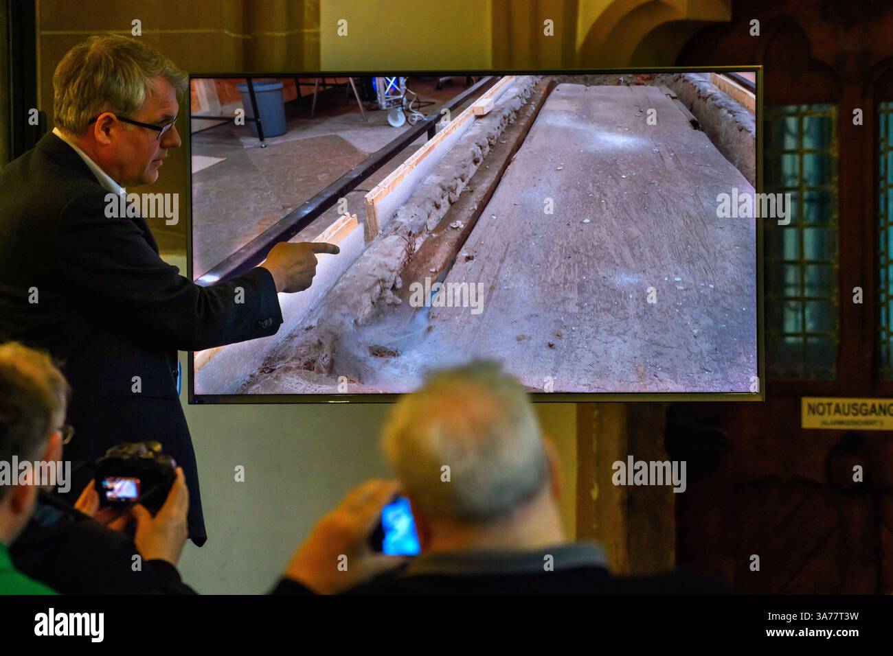 26. März 2025, Sachsen-Anhalt, Magdeburg: In der Sakristei des Magdeburger Doms erklärt Donat Wehner (l) vom Landesamt für Denkmalpflege und Archäologie Sachsen-Anhalt Medienvertretern die Beschädigung der Holzkiste im offenen Sarkophag mit Hilfe einer Live-Videoübertragung aus dem Gehege rund um das Grab Ottos des Großen. Seit Januar führen das Landesamt für Denkmalpflege und Archäologie und die Kulturstiftung Sachsen-Anhalt Untersuchungen zur Erhaltung des Denkmals durch. Die Ergebnisse der Untersuchungen bis Stockfoto
