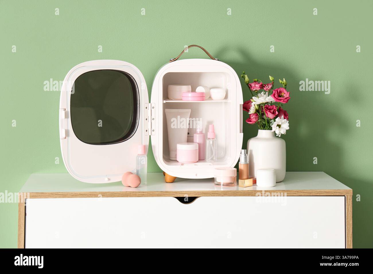 Öffnen Sie den kleinen Kühlschrank mit Kosmetikprodukten und Blumen in der Vase auf der Kommode vor grünem Hintergrund. Nahaufnahme Stockfoto