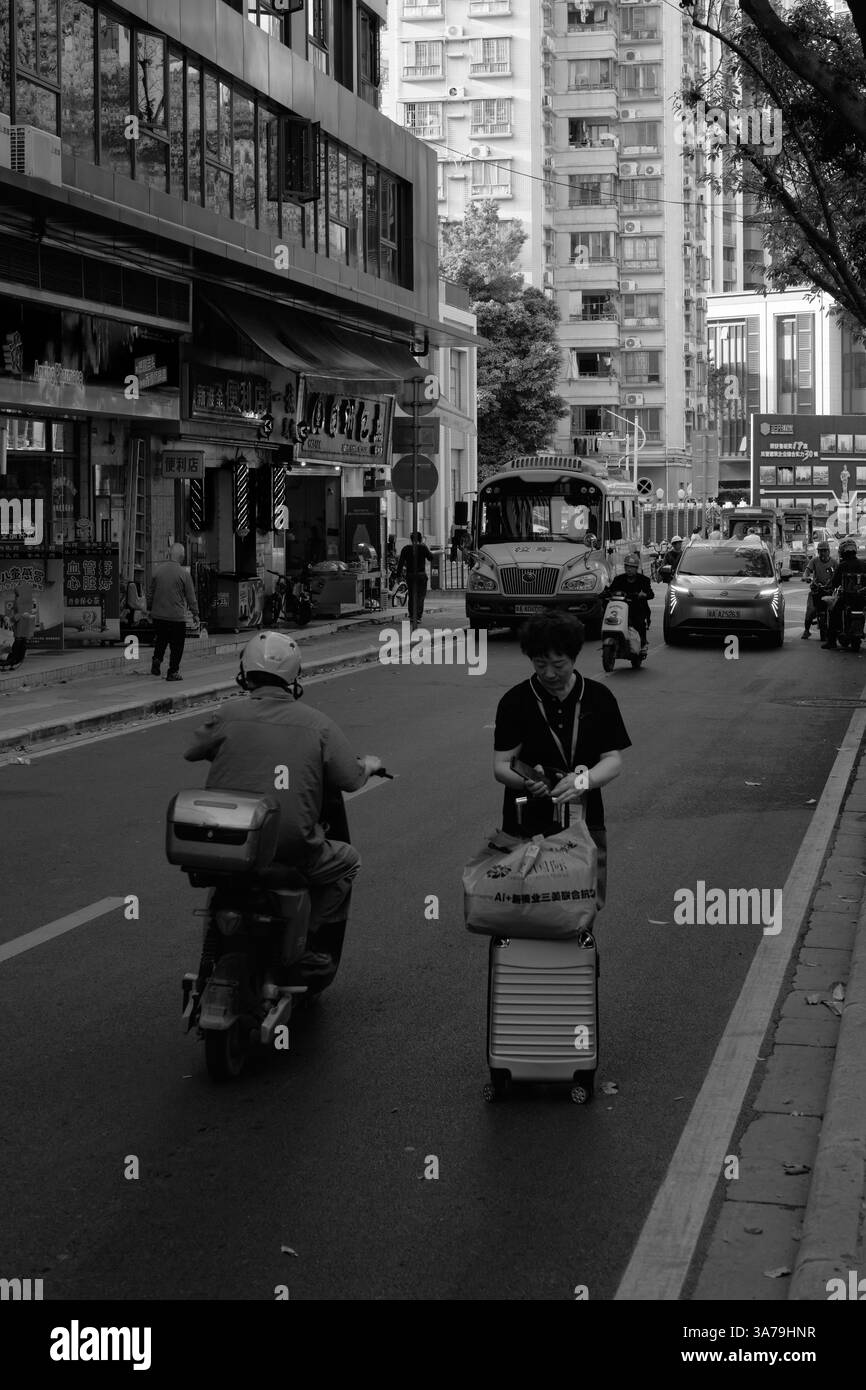 Eine Frau mit einem Koffer steht auf einer geschäftigen Straße in Guangzhou, China, während Autos vorbeifahren. Ein monochromes Stadtbild, das das Leben und die Bewegung der Stadt einfängt. Stockfoto