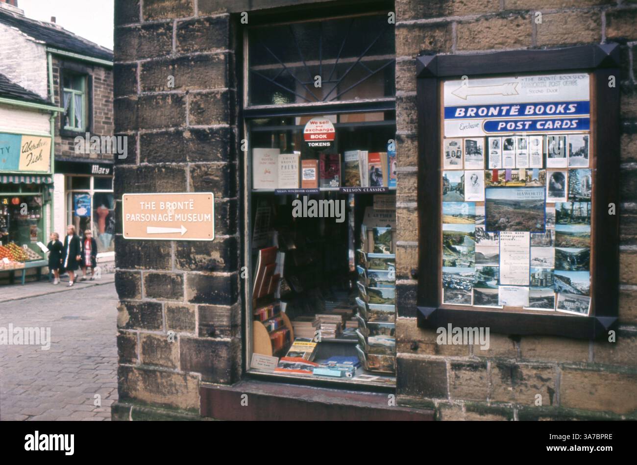 Eine Straßenszene aus den 1960er Jahren vor dem Brontë Bookshop and Post Office in Haworth, Yorkshire. Mit einer bezaubernden Ausstellung von klassischen Büchern, Postkarten und Schildern, die auf das Pfarrhaus von Brontë zeigen. Stockfoto
