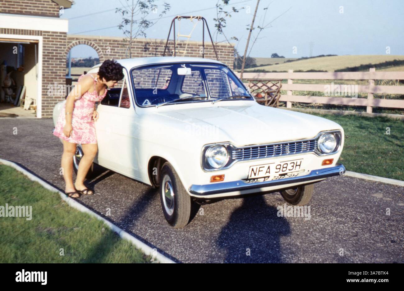 Eine Frau inspiziert einen weißen Ford Escort Mk1, der in den späten 1960er oder frühen 1970er Jahren auf einer Einfahrt auf dem britischen Land geparkt wurde Das Fahrzeug mit H-Schild (1969–1970) Stockfoto