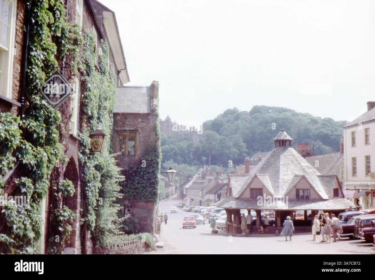 Die Dunster High Street in Somerset ist eine lebendige Szene aus den 1960er-Jahren mit dem legendären achteckigen Garnmarkt im Zentrum. Einheimische und Besucher treffen sich in der Nähe von Oldtimern, während Evy die Mauern des historischen Luttrell Arms Hotel auf der linken Seite erklimmt. Stockfoto