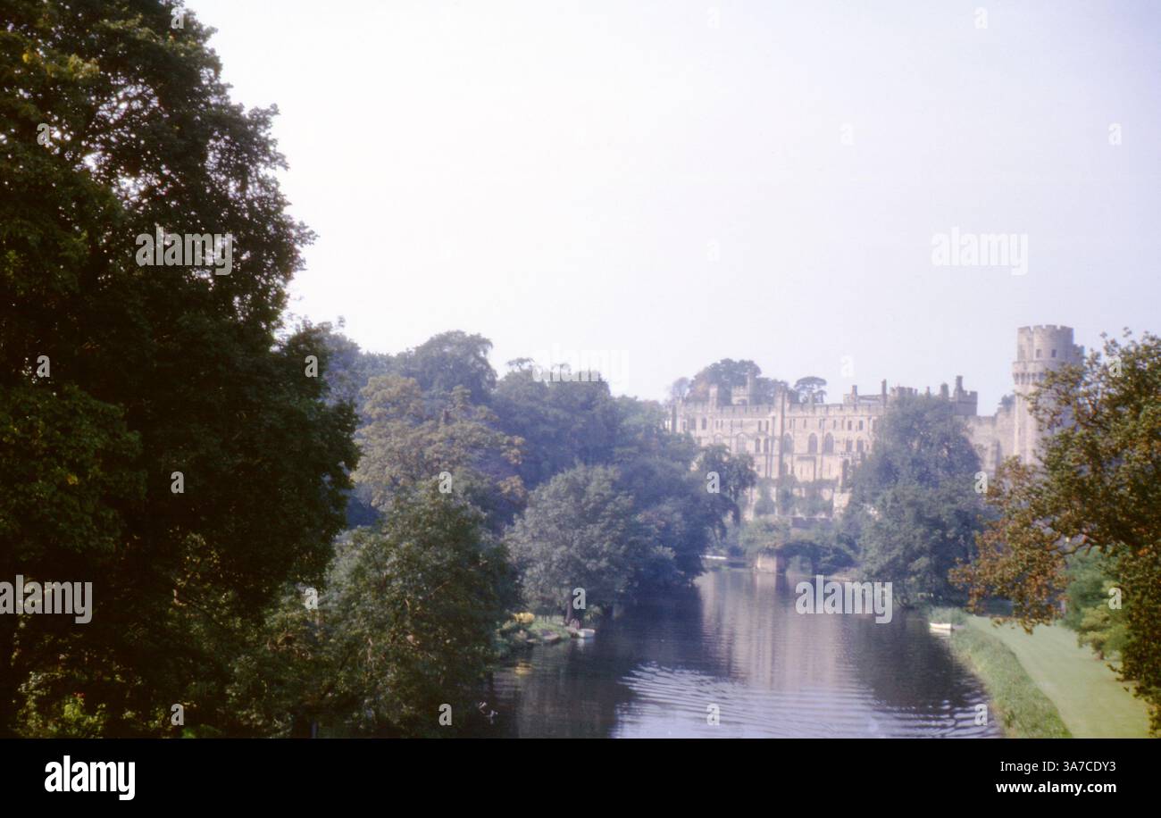 Warwick Castle von der anderen Seite des Flusses Avon, umgeben von alten Bäumen und spiegeln sich im Wasser. Eine friedliche Szene, in der die Pracht der mittelalterlichen Festung an einem trüben Tag in den 1960er Jahren festgehalten wird Stockfoto