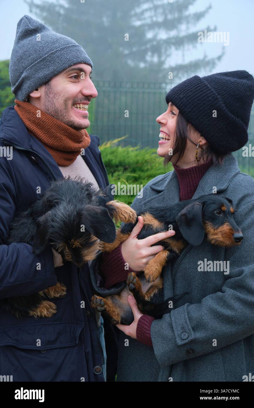 Junge Paare lachen und halten ihre beiden liebenswerten Hunde in einem nebeligen Park an einem kalten Wintertag Stockfoto
