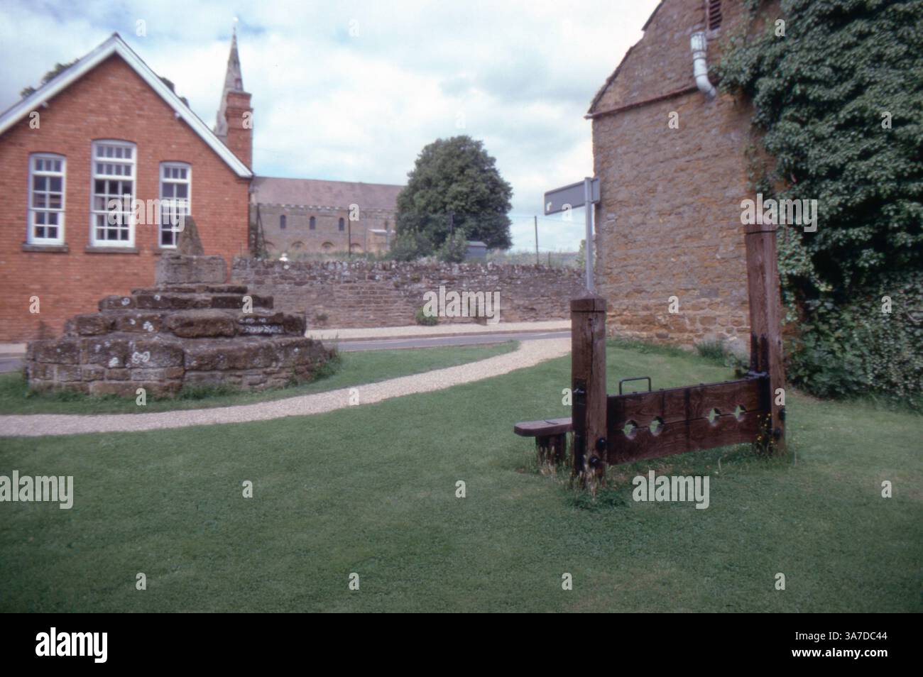 Historische Bestände und eine mittelalterliche Dorfkreuzung in Brixworth, Northamptonshire, England, fotografiert im Jahr 1993. Die Vorräte, die einst zur öffentlichen Bestrafung verwendet wurden, stehen auf einem grasbewachsenen Gebiet in der Nähe der Pfarrkirche und der alten Dorfgebäude. Stockfoto