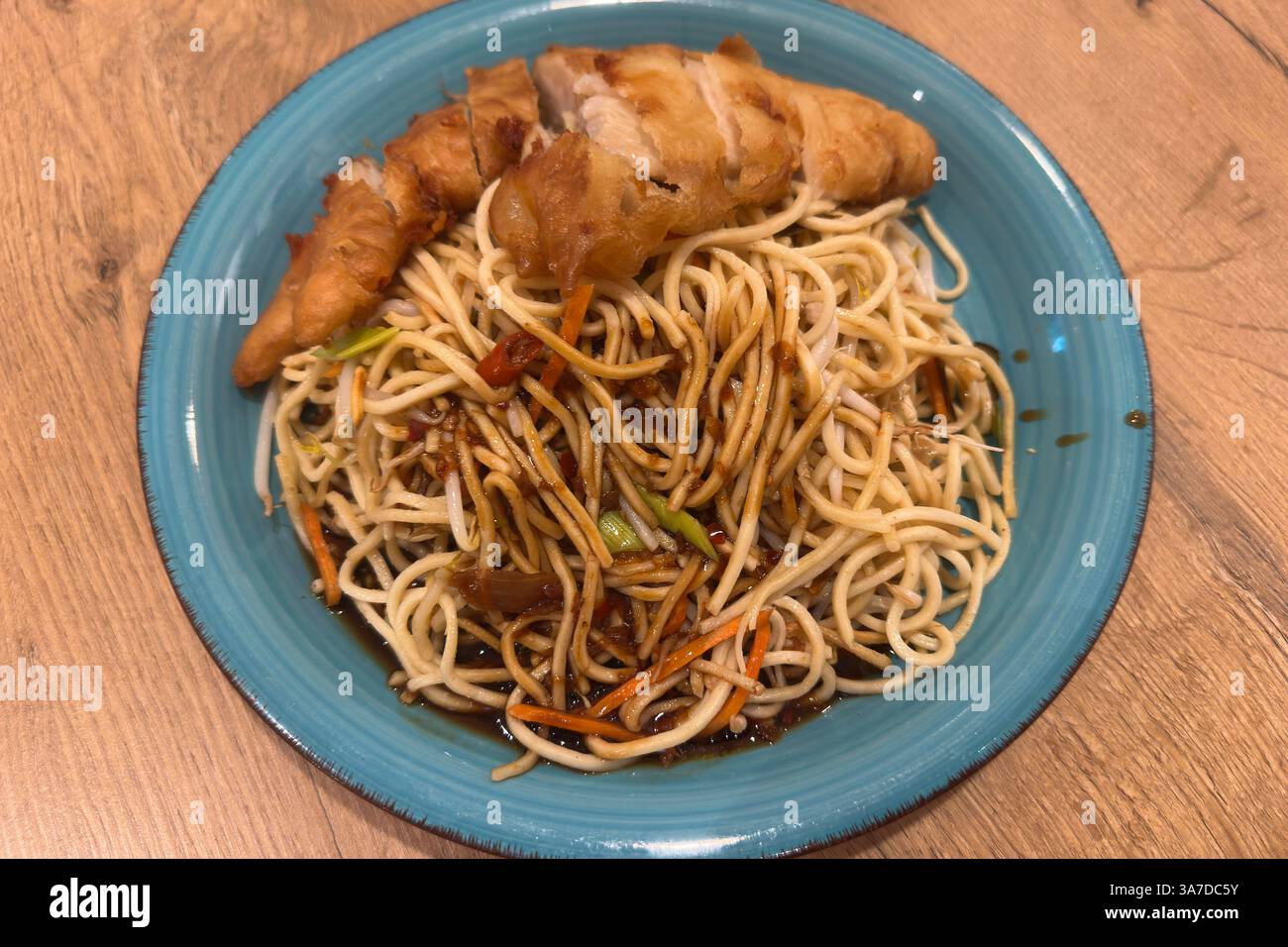 , Deutschland, Rheinland-Pfalz, 27.03.2025, frisch gebratene Nudeln mit Gemüse, Sojasauce und knusprigem Hähnchenfilet – serviert auf blauem Teller. Stockfoto
