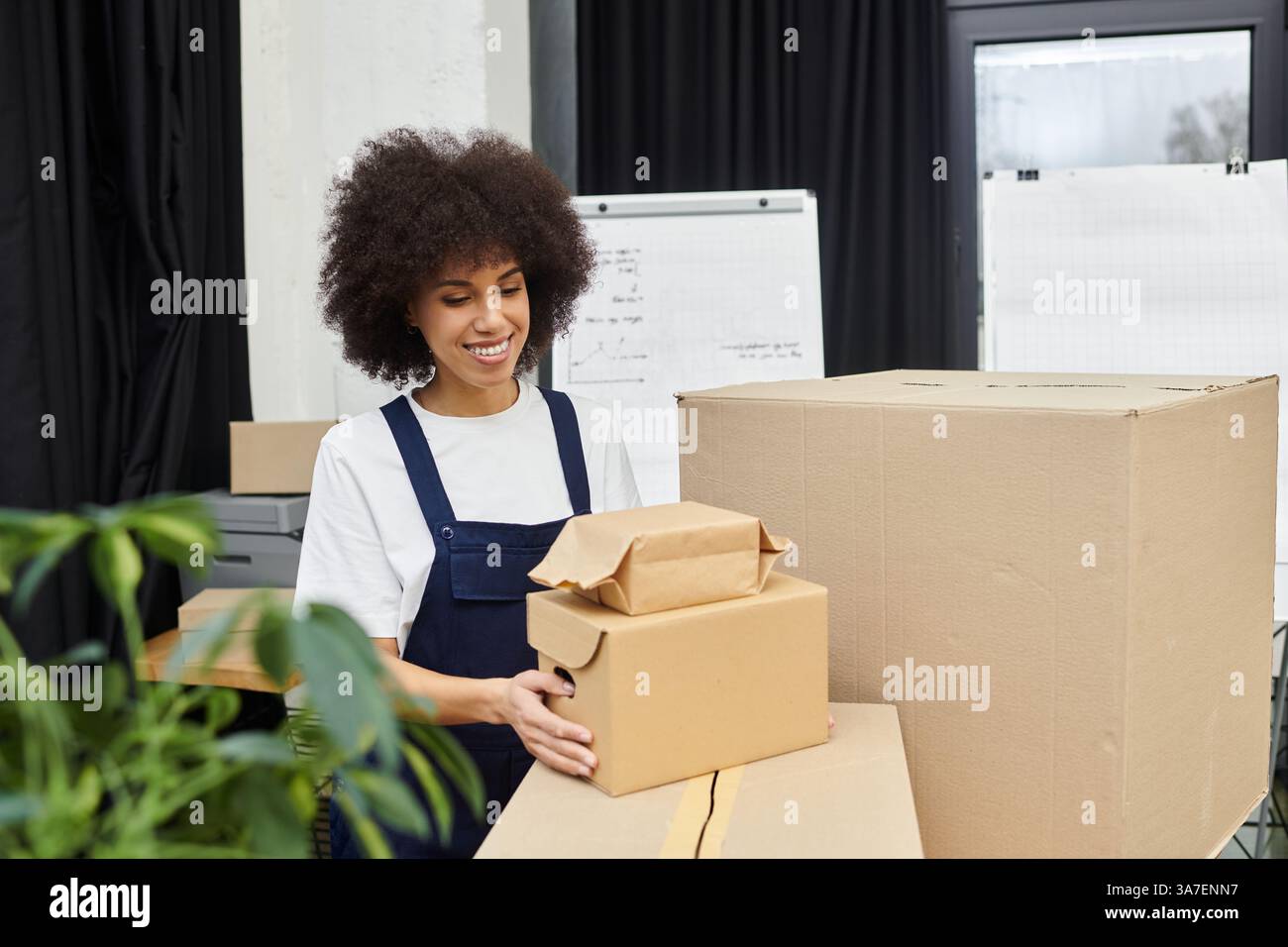 Eine junge Frau lächelt, während sie Kisten und Pakete in einer modernen Büroumgebung organisiert. Stockfoto