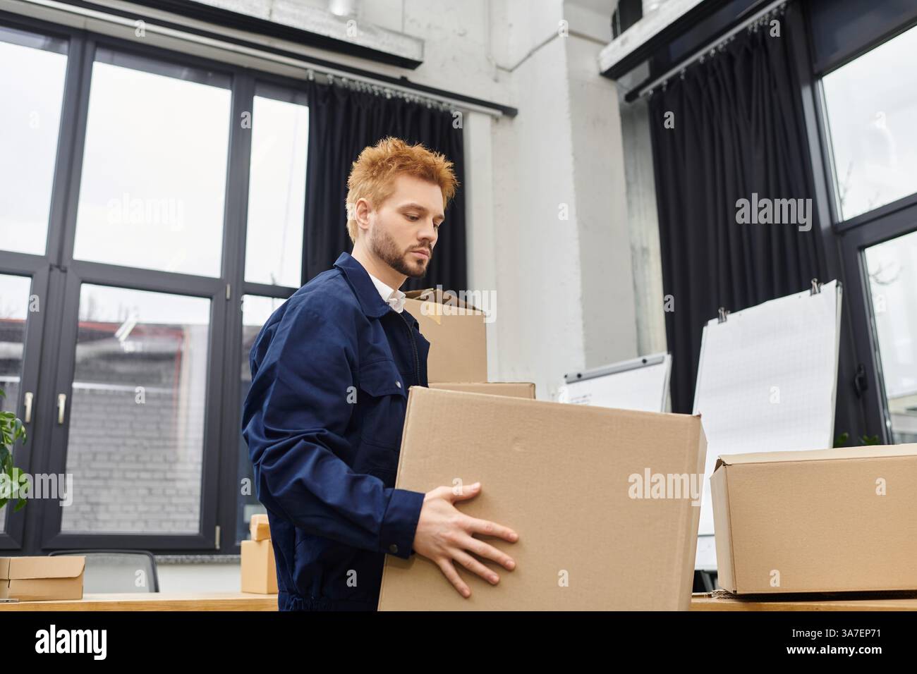 Ein Mann konzentriert sich intensiv, während er eine Pappschachtel in einer hellen, modernen Büroumgebung hebt. Stockfoto