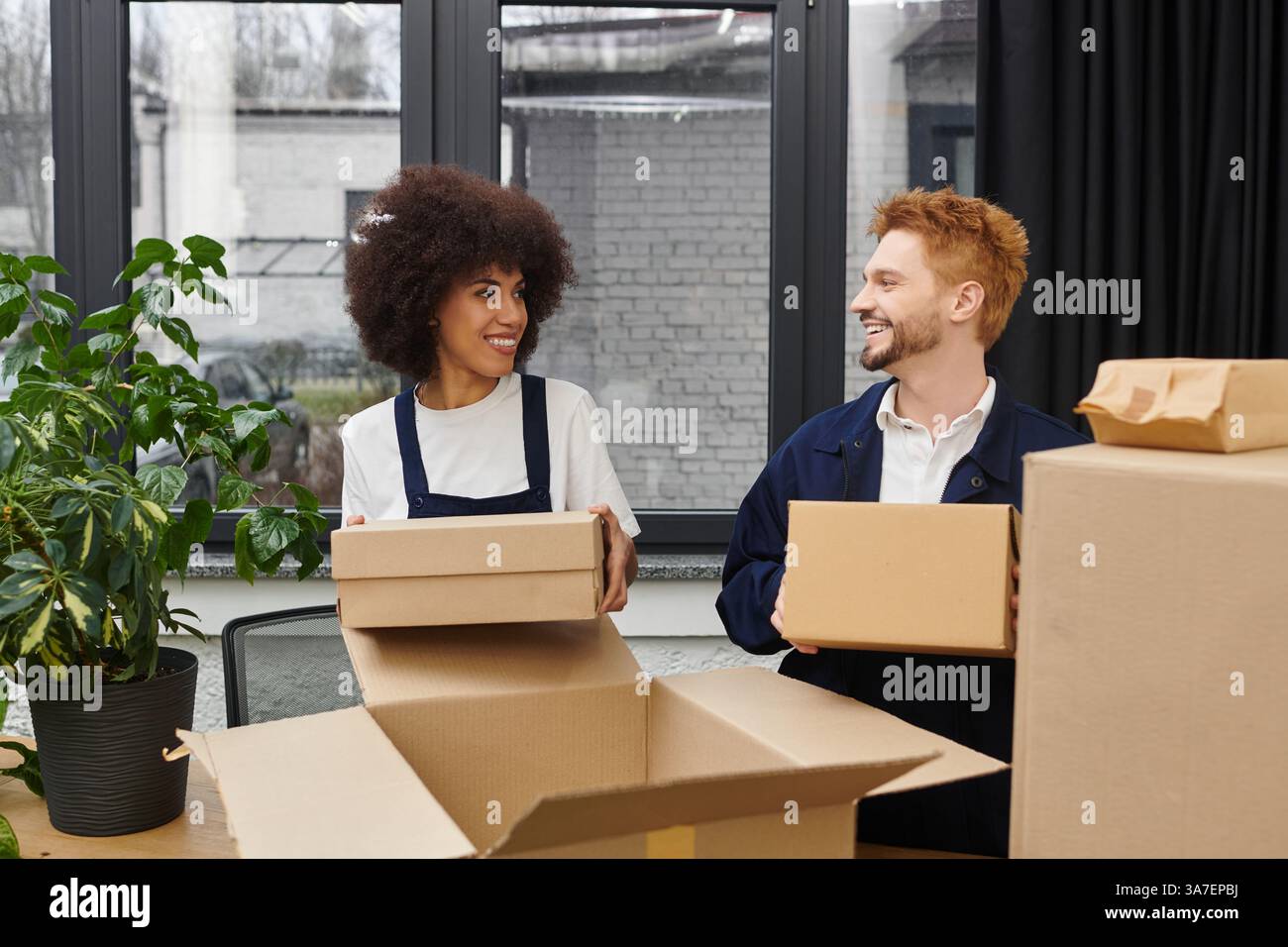 Zwei Leute sortieren freudig Umzugskästen in einem modernen Büro mit nahegelegenen Pflanzen. Stockfoto