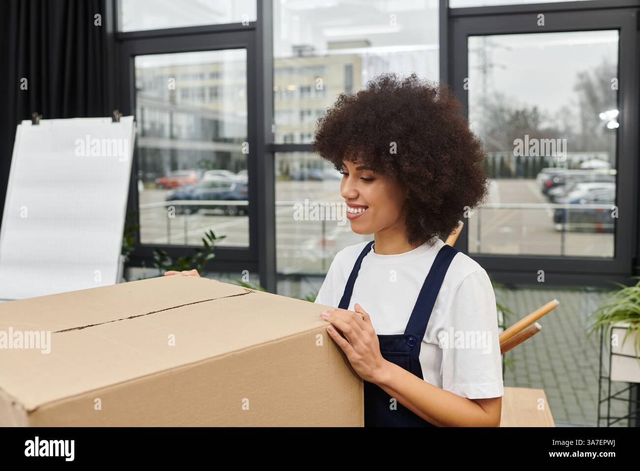 Eine Frau lächelt, während sie eine große Kiste in einem modernen Büro auspackt, voller Aufregung. Stockfoto