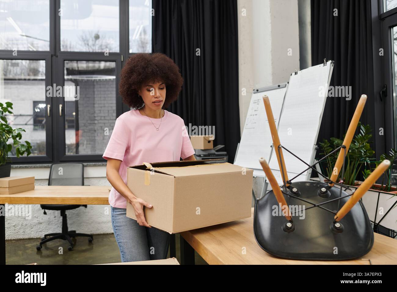 Eine junge Frau packt eine Kiste zwischen Möbeln und beweglichen Vorräten in einem modernen Büro. Stockfoto
