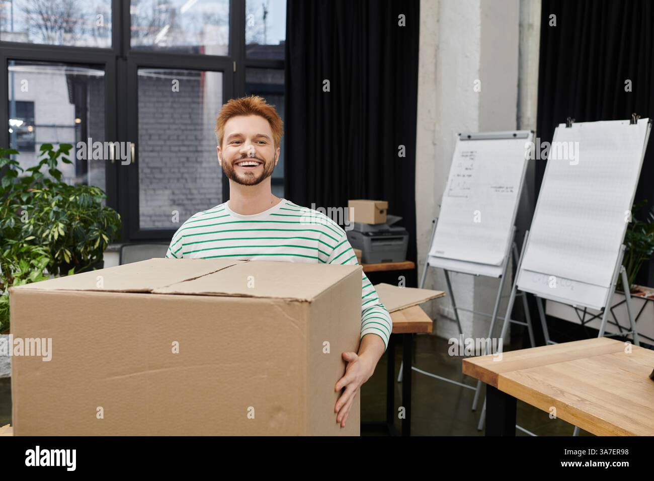Happy man trägt eine große Kiste, während er seinen Arbeitsbereich für einen Umzug organisiert. Stockfoto