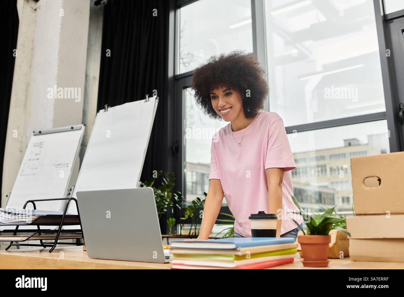 Eine Frau in einem hellrosafarbenen Hemd konzentriert sich auf ihren Laptop, umgeben von Verpackungen und Pflanzen. Stockfoto