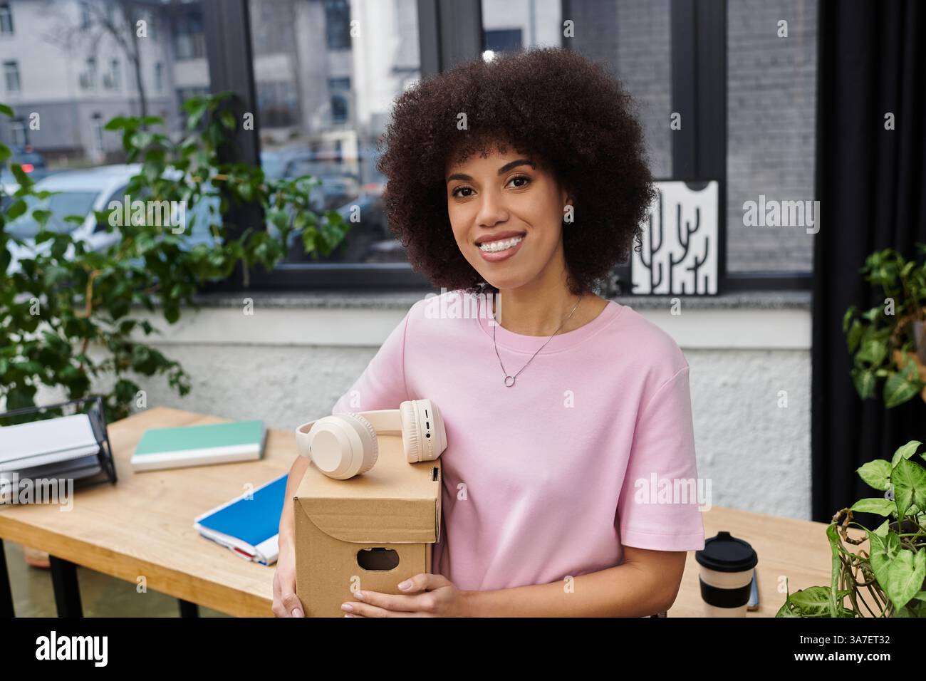 Eine fröhliche Frau hält eine Pappschachtel und Verpackungsmaterialien, während sie von Pflanzen umgeben ist. Stockfoto