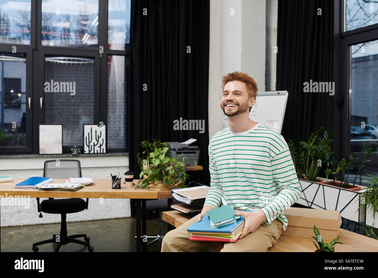 Ein fröhlicher Mann arrangiert ein stilvolles Büro mit einem Stapel bunter Bücher. Stockfoto