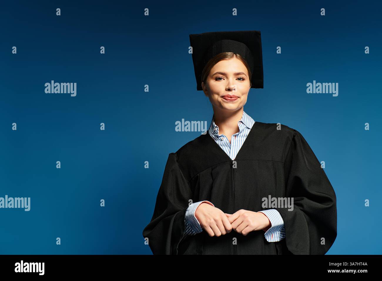 Die fröhliche junge Frau steht stolz mit einer Abschlussmütze und einem Kleid vor blauem Hintergrund. Stockfoto