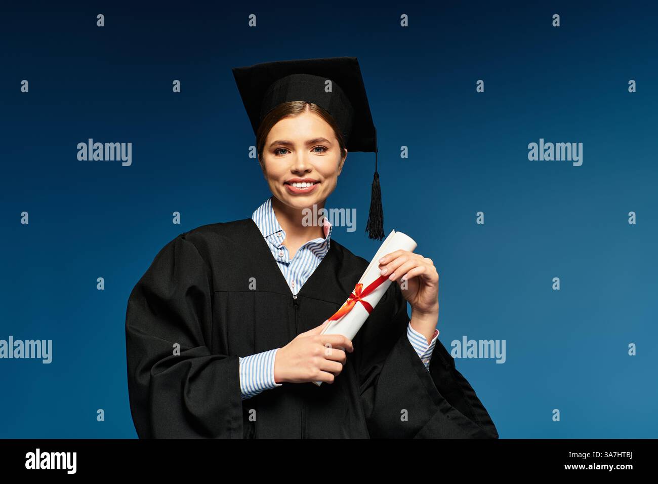 Eine junge Frau in Abschlusskleidung, die stolz ihr Diplom vor blauem Hintergrund hält. Stockfoto