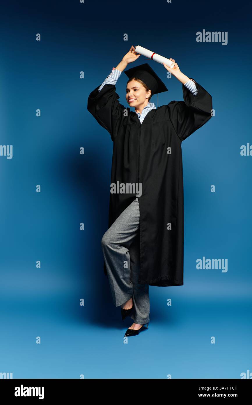 Eine glückliche junge Frau hebt ihr Diplom an, während sie ein Abschlusskleid trägt und ihren Erfolg zeigt. Stockfoto