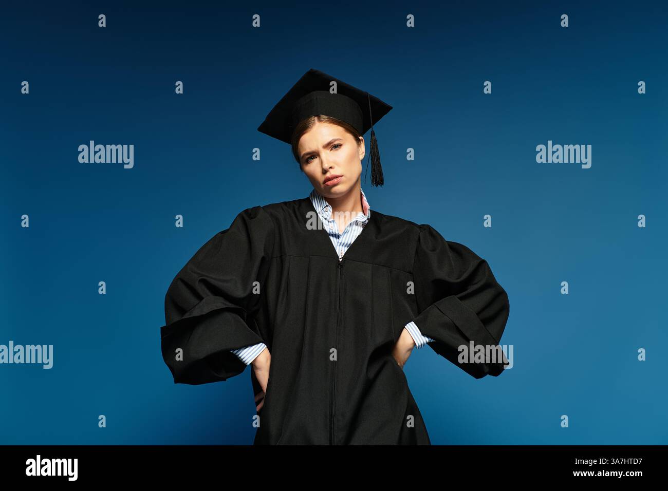 Eine stolze junge Frau in Abschlusskleidung steht selbstbewusst vor einem kühnen blauen Hintergrund. Stockfoto