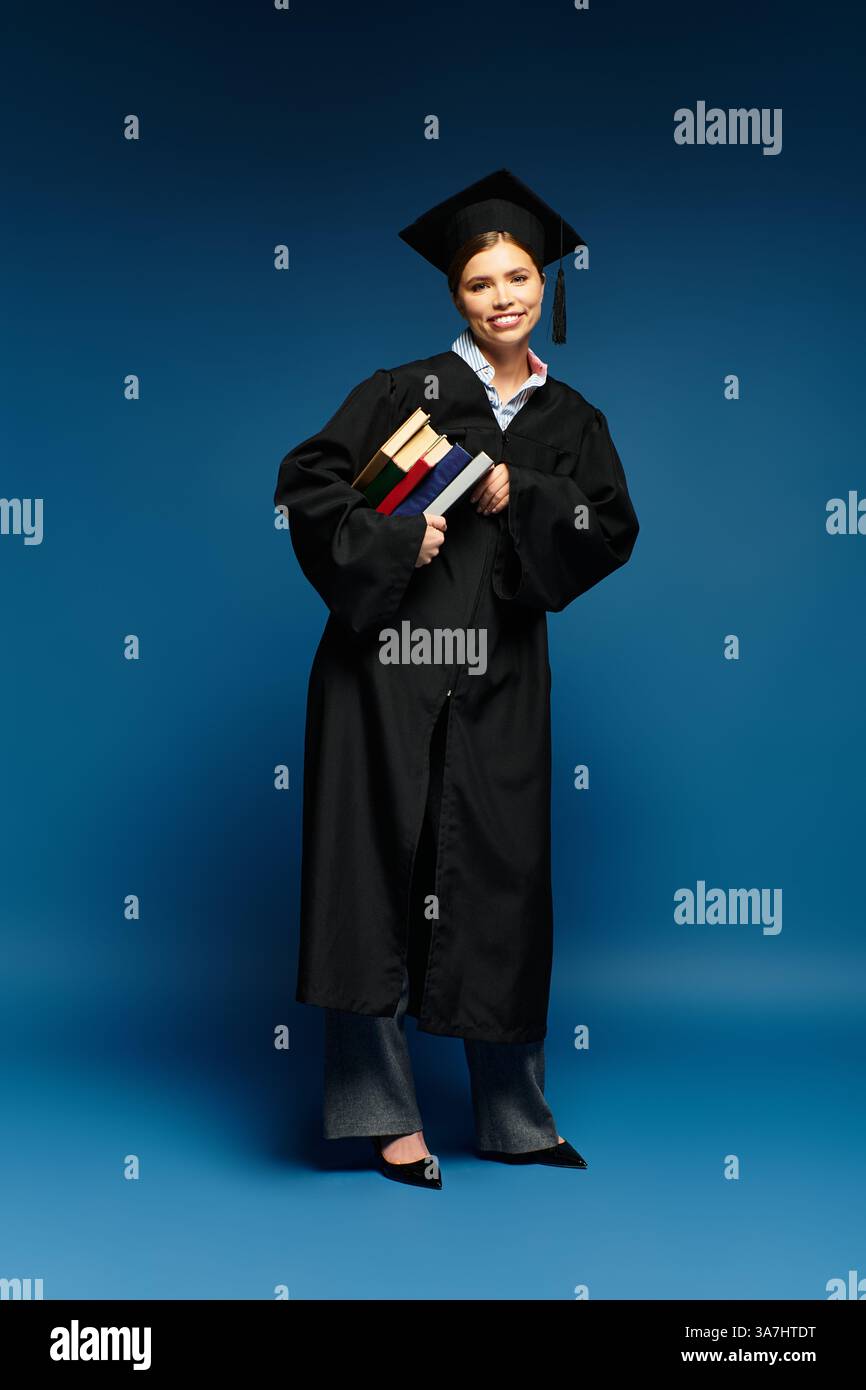 Eine junge Frau im Abschlusskleid und Mütze hält Bücher und lächelt vor blauem Hintergrund. Stockfoto