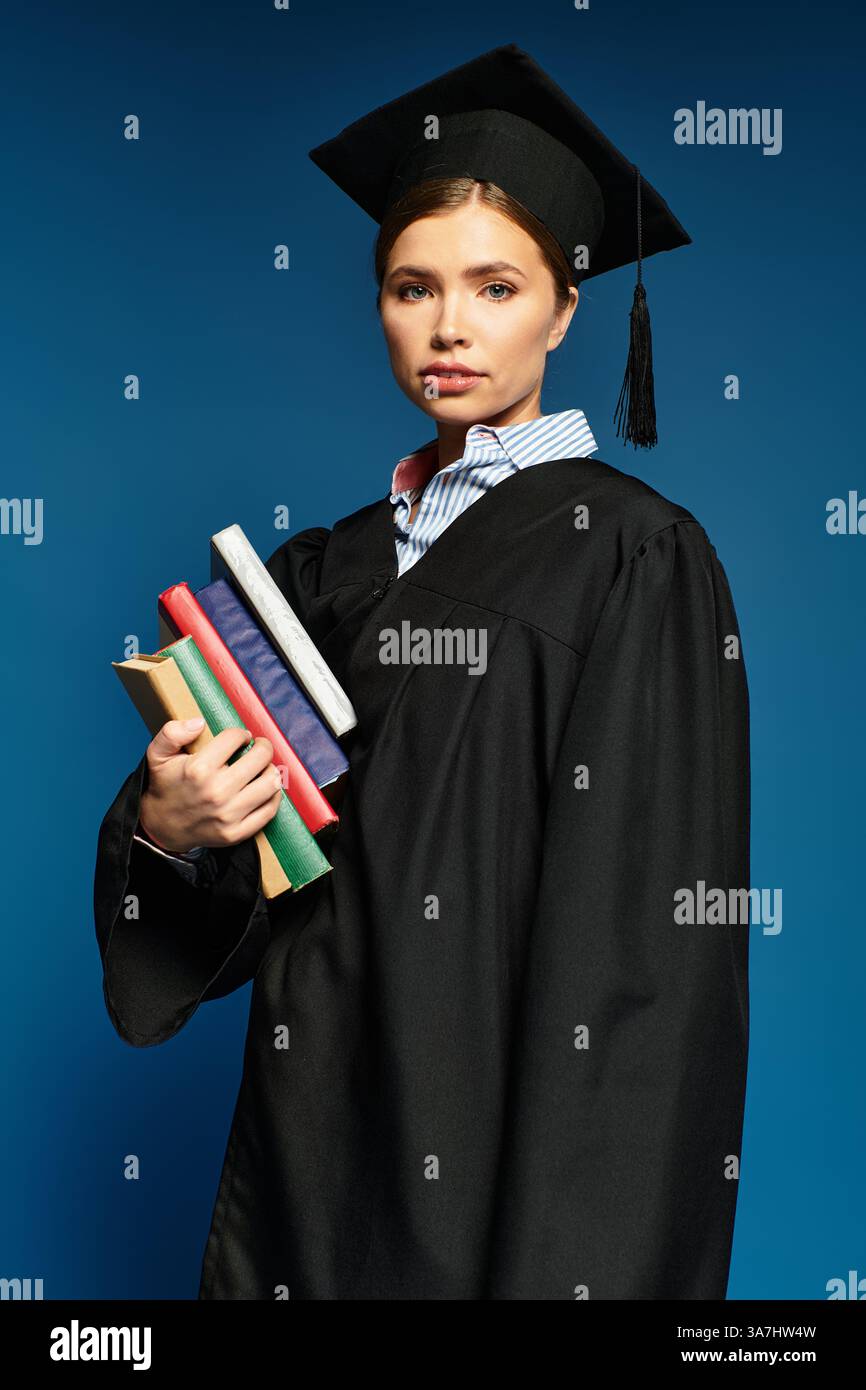 Eine stolze junge Frau im Abschlusskleid hält Bücher und strahlt vor blauem Hintergrund. Stockfoto