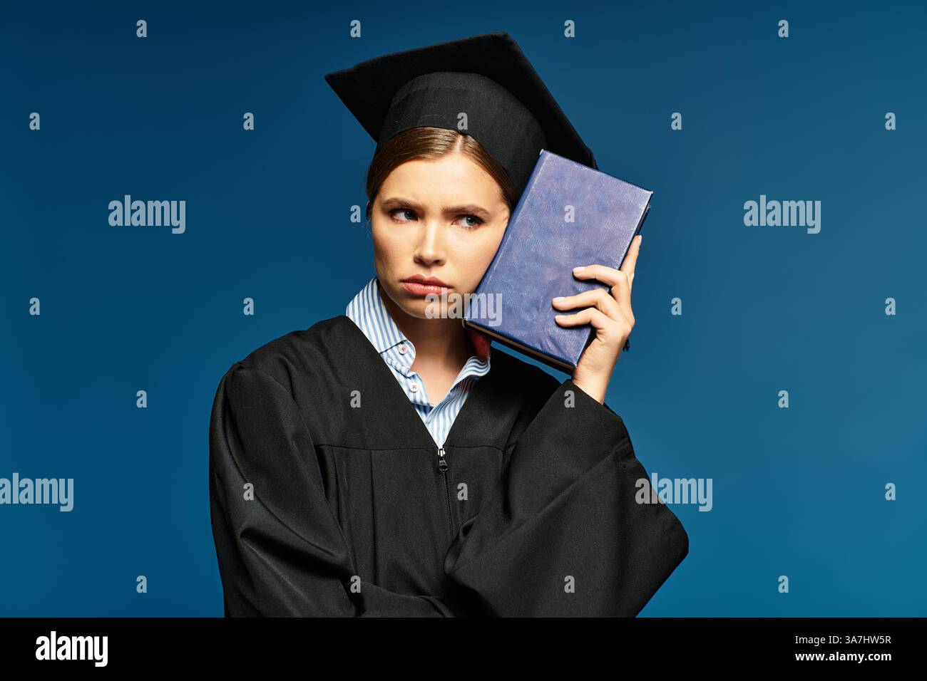 Eine nachdenkliche junge Frau in Abschlusskleidung hält ihr ein Buch an den Kopf. Stockfoto