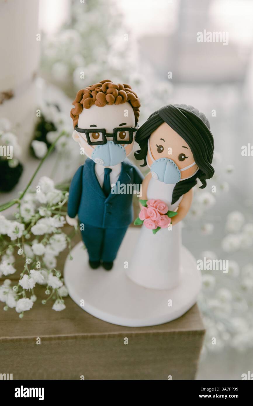 Der einzigartige Hochzeitstortendeckel zeigt ein maskiertes Paar mit zarten Blumen, die Liebe inmitten von Herausforderungen symbolisieren. Stockfoto