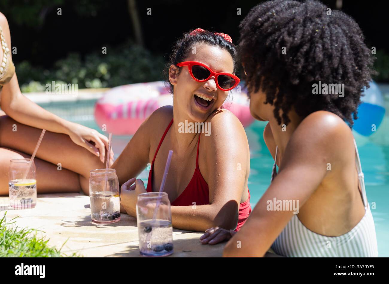 Lachende Freundinnen, die an sonnigen Tagen erfrischende Getränke am Pool genießen, Kopierraum Stockfoto
