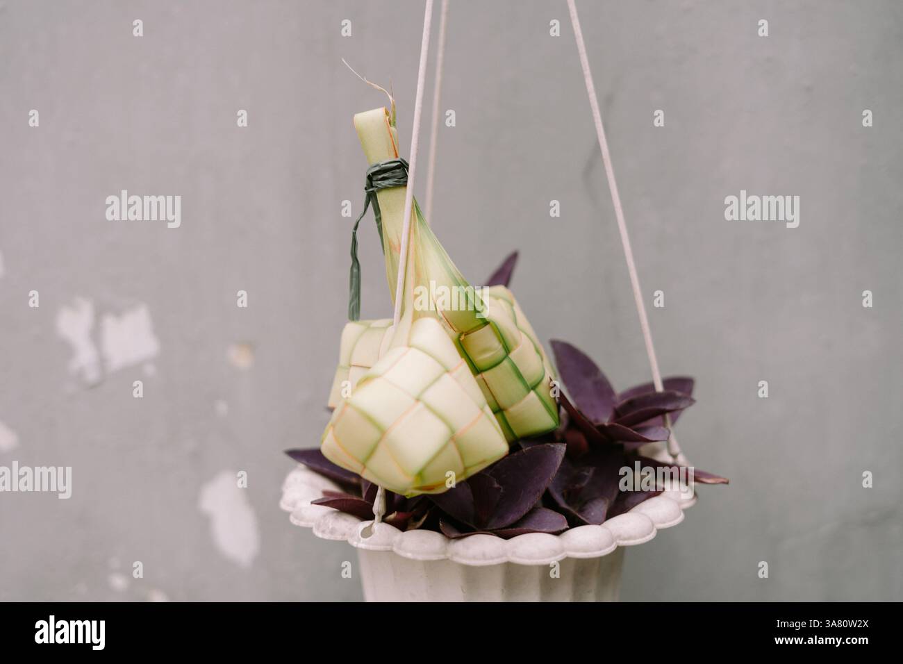 Nahaufnahme von Ketupat, einem traditionellen Reisknödel, der bei festlichen Feierlichkeiten in südostasiatischen Ländern beliebt ist und über einem Topf mit violetter Blüte hängt Stockfoto