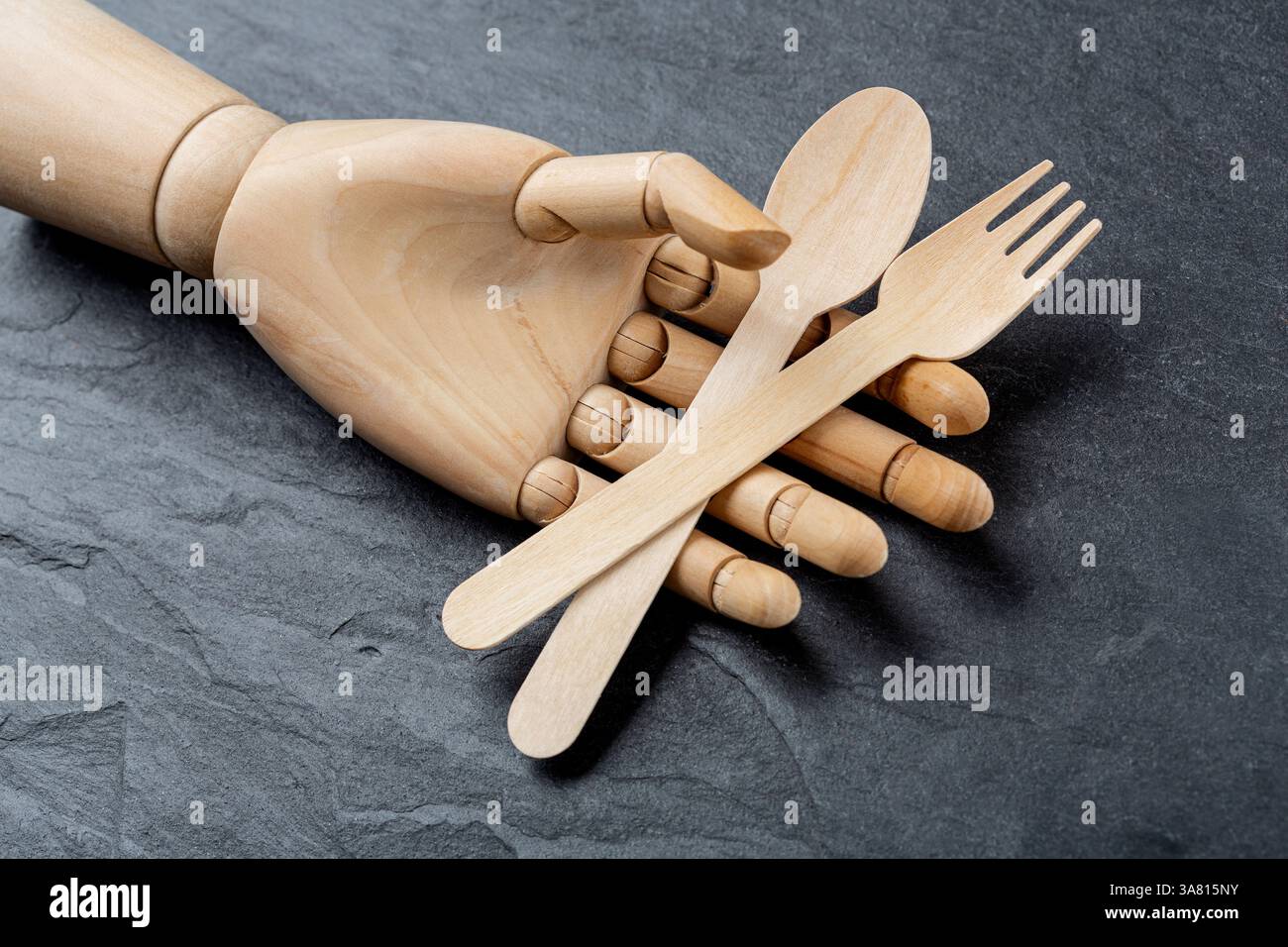 Hölzerner Einweglöffel und Gabel in einer hölzernen Hand einer Schaufensterpuppe auf schwarzem Schiefergrund. Gesundes Lebensmittelkonzept und Naturproduktkonzept Stockfoto