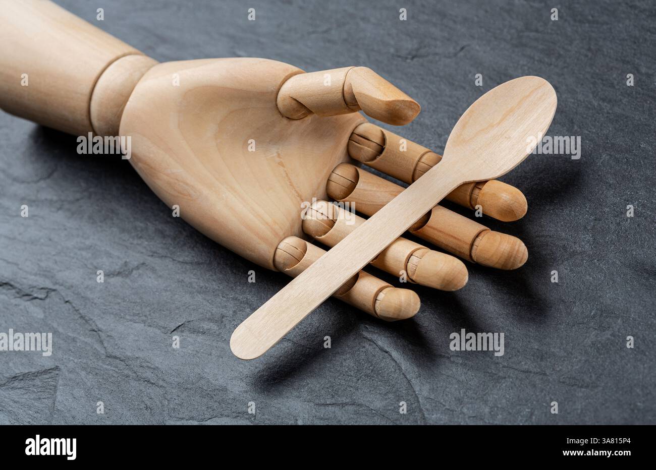 Hölzerner Einweglöffel in einer hölzernen Hand einer Schaufensterpuppe auf schwarzem Schiefergrund. Gesundes Lebensmittelkonzept und Naturproduktkonzept Stockfoto