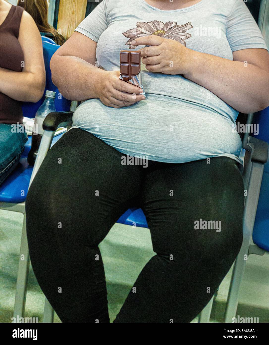 Fettleibige weiße Frau, die in einem Wartezimmer des Krankenhauses saß und eine Tafel Schokolade hielt, mit ihrem Gesicht nicht sichtbar im Rahmen Stockfoto